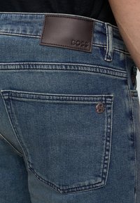 Blaue Jeans aus Denim mit einem Lederpatch mit der Marke "BOSS" über der Gesäßtasche und einem kleinen Metalllogo.