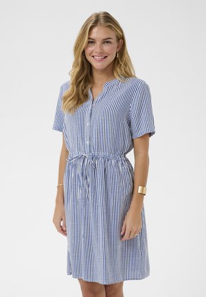 Femme aux cheveux blonds souriante, portant une robe à manches courtes à rayures verticales bleues et blanches avec des boutons et une taille à cordon, accessoires dorés.