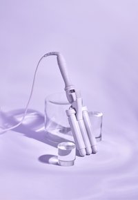 Fer à friser blanc avec trois cylindres, poignée texturée et affichage numérique. Présenté sur un fond violet doux avec des objets en verre clair.