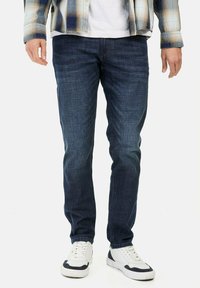 Jean en denim bleu foncé avec une coupe slim, texture lisse, poches standard et finition délavée, associés à des baskets blanches.
