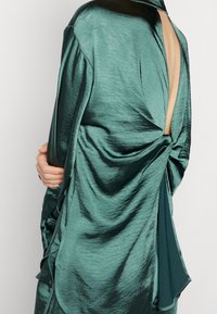 Vestido de satén en color verde azulado con un diseño drapeado, que presenta un corte profundo en la espalda y detalles anudados en la cintura. Textura suave con un acabado brillante.