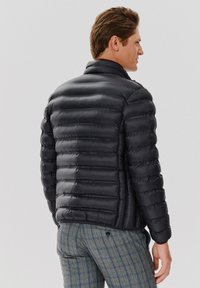 Czarna kurtka puchowa o wyraźnej konstrukcji, poziome pikowanie, wysoki kołnierz oraz gładka, błyszcząca tekstura. Połączona z materiałowymi spodniami w kratę.