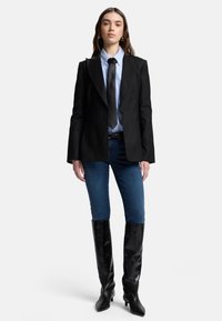 Svart skräddarsydd kavaj, ljusblå skjorta med krage, svart slips, blå denimjeans och knähöga svarta läderstövlar med spetsiga tår.