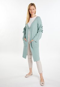 Cardigan color turchese chiaro con maniche arricciate, scollatura a V profonda e chiusura frontale con bottoni. Include due tasche frontali ed è abbinato a pantaloni beige.