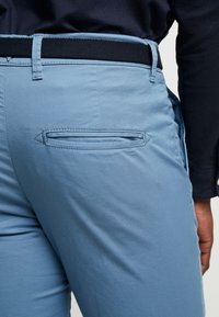 Světlounce modré kalhoty z texturované látky, černý tkaný pás a jedna šikmá zadní kapsa. Bezvráskový vzhled s hladkým prošitím.