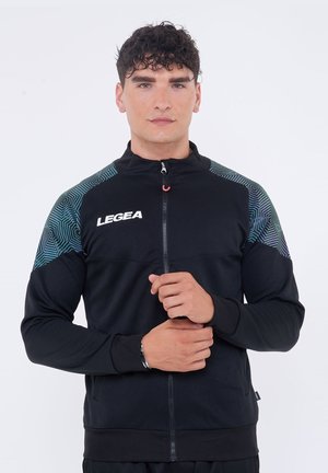 Uomo che indossa una giacca sportiva nera con zip, con motivi geometrici blu sulle spalle e il logo "LEGEA" sul petto, che sistema il polsino della manica.