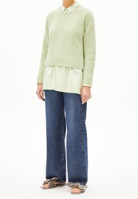 Vihreä ribbikuvioinen neule, kerrostettuna vaaleanvihreän paidan päälle, yhdistettynä leveälahkeisiin tummansinisiin farkkuihin ja harmaisiin liukuisiin sandaaleihin, joissa on solmittuja yksityiskohtia.