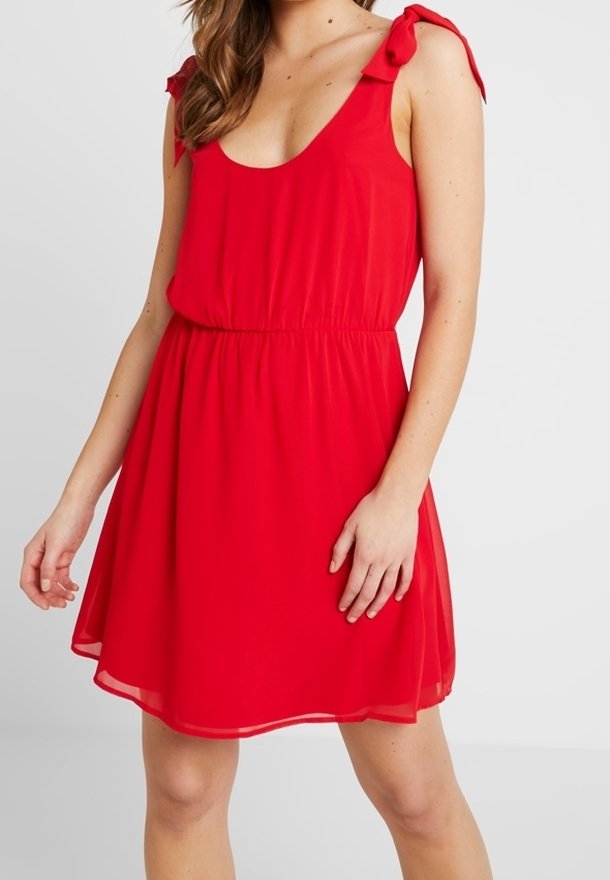 Femme portant une robe rouge sans manches avec des bretelles nouées aux épaules et une taille élastique, debout devant un fond clair uni.