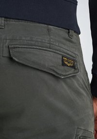 Detailní záběr na kapsu olivově zelených cargo kalhot s klopou a černou nášivkou PME Legend American Classic, nošenou s tmavým ribbovaným svetrm.