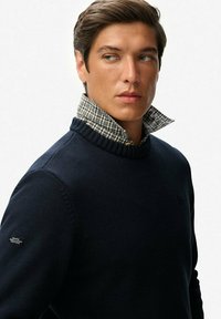 Pull en mailles bleu marine avec un col côtelé, superposé sur un col de chemise à carreaux. Présente un petit logo sur la manche gauche.