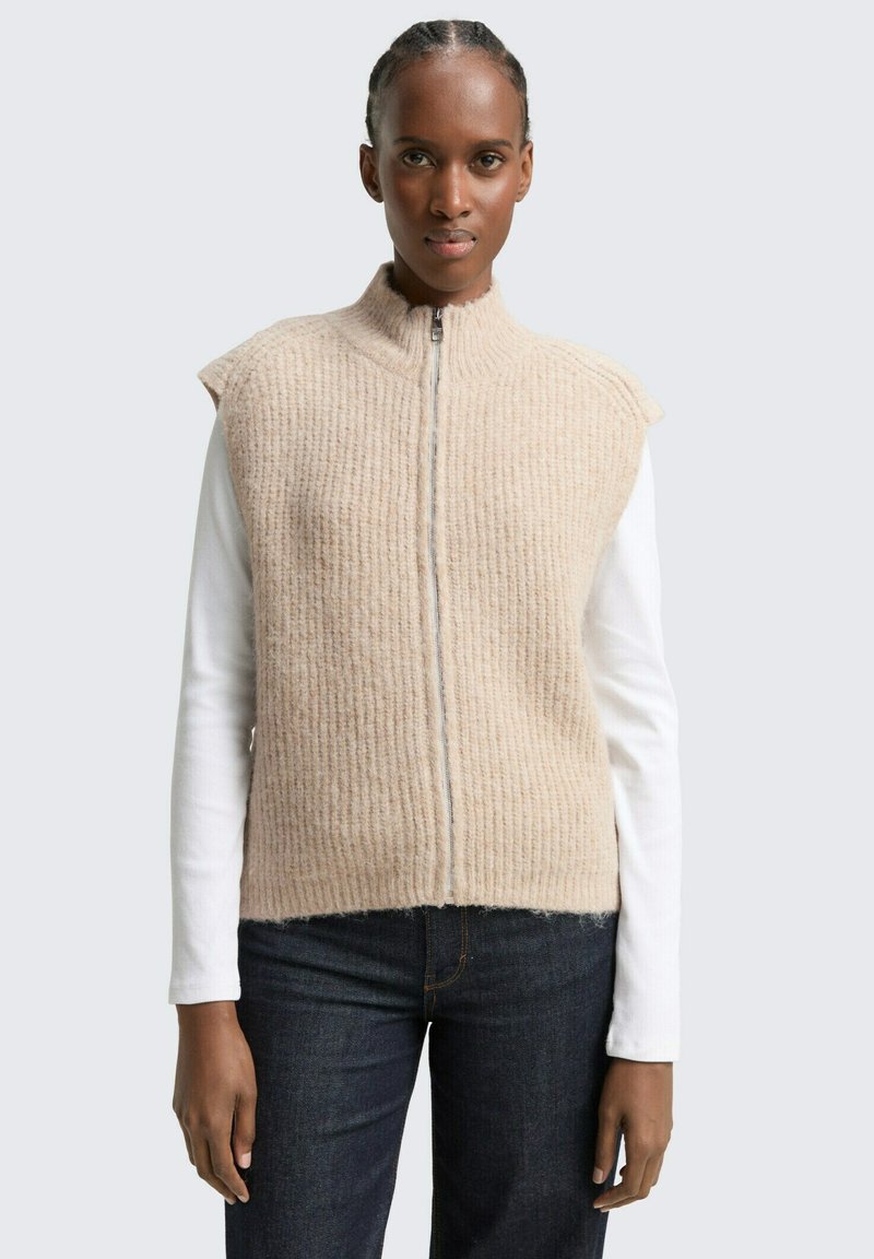 Beige wollen vest met ribbeltextuur, rits aan de voorkant en mouwloos ontwerp, gedragen over een wit overhemd met lange mouwen, gecombineerd met donkere jeans.