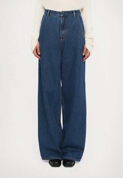 Brede, donkerblauwe jeans met een hoge taille, gecombineerd met zwarte schoenen en een crèmekleurig, geribbeld langsliefs T-shirt met verlengde manchetten.