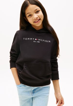 Sudadera negra con logo de "TOMMY HILFIGER" y fecha de establecimiento, con un bolsillo frontal y puños acanalados, combinada con jeans de color azul claro.