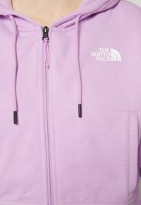 Sweat à capuche violet clair à zip avec cordons de serrage et logo "The North Face" en blanc sur la poitrine gauche.