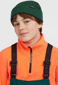 Groene geribbelde beanie, oranje fleece met halve ritssluiting, en verstelbare zwarte banden met logo, gecombineerd met een donker groene bretellenoverall.