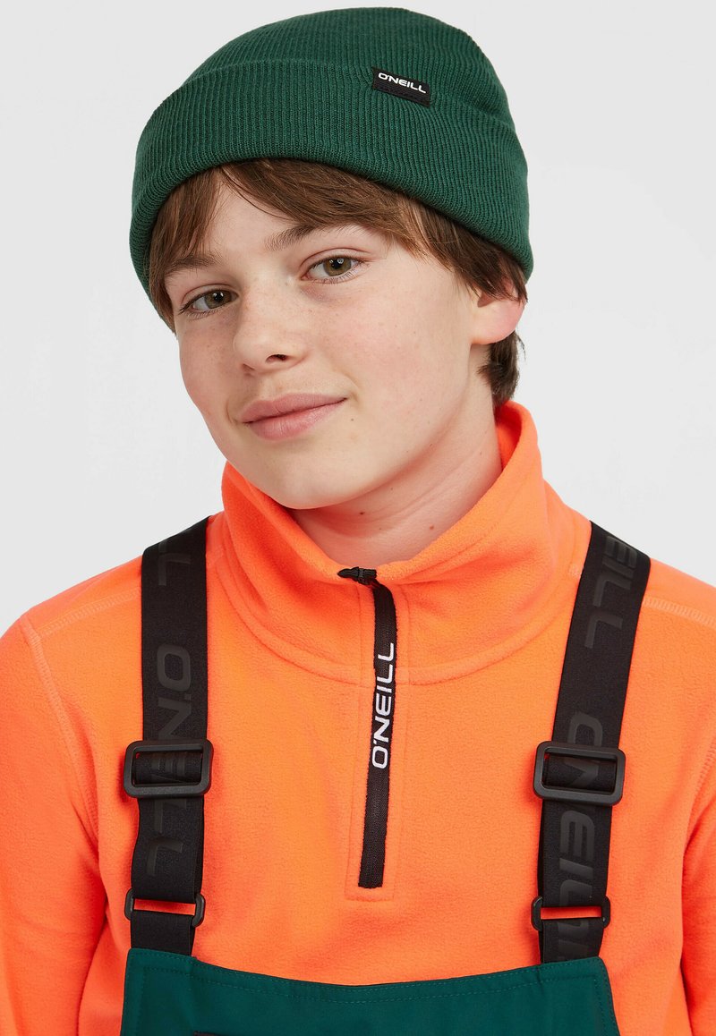 Groene geribbelde beanie, oranje fleece met halve ritssluiting, en verstelbare zwarte banden met logo, gecombineerd met een donker groene bretellenoverall.