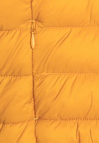 Veste matelassée jaune avec une texture capitonnée, présentant des coutures marquées et une fermeture éclair centrale avec un tirette rond.