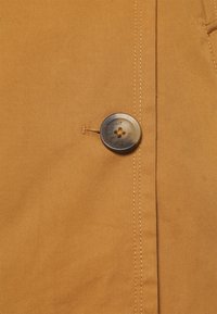 Tissu marron en gros plan avec un bouton gris à quatre trous cousu et des coutures visibles le long du bord.