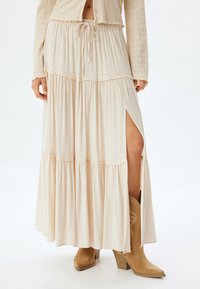Jupe maxi beige clair à étages avec taille à cordon de serrage et fente sur le côté, présentant une texture douce et plissée ainsi que des détails de couture décoratifs.