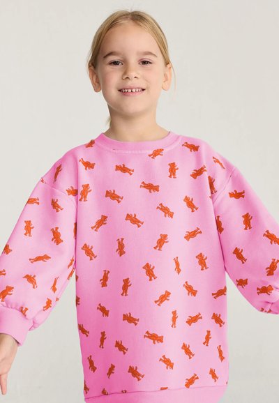Jeune fille aux cheveux blonds portant un sweat-shirt rose couvert de petites illustrations d'ours orange, souriante sur un fond clair.