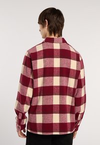 Mand, der står med ryggen til, iført en langærmet rød og beige ternet flannel skjorte med krave og knaplukninger ved håndleddene.