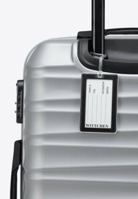 Valigia rigida d'argento con finitura testurizzata, manico nero e etichetta identificativa attaccata che mostra i campi di contatto. Presenta un design elegante e aerodinamico.