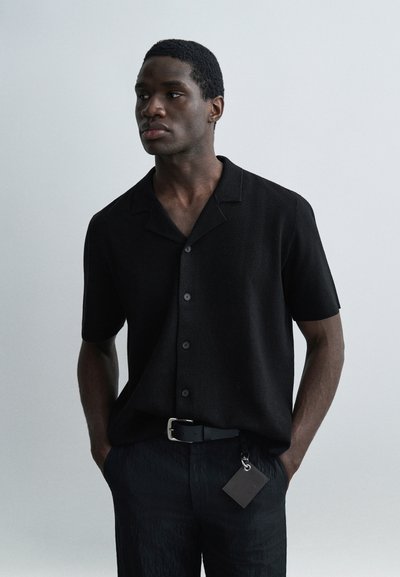 Homme aux cheveux courts portant une chemise noire à manches courtes boutonnée et un pantalon noir, les mains dans les poches, sur fond gris.
