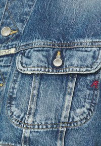 Blå denimjacka med metallknappar, en bröstficka och kontrasterande orange sömmar. Strukturerad tyg med lätt bleknade detaljer.