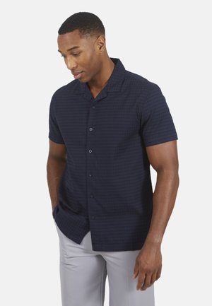 Mann trägt ein marineblaues kurzärmliges Hemd mit Knöpfen und hellgraue Hose, steht mit den Händen in den Taschen und blickt nach unten.