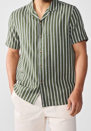 Mężczyzna w oliwkowej koszuli na krótki rękaw z białymi pionowymi paskami i jasnobeżowych spodniach, stojący z jedną ręką w kieszeni.