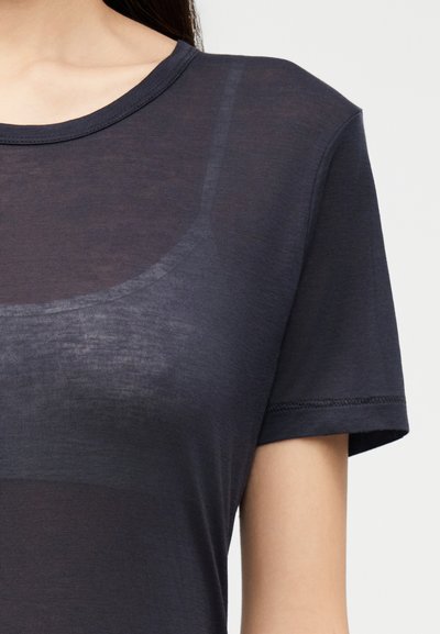 Vrouw draagt een donker, semi-transparant T-shirt met korte mouwen over een zwart ondershirt, afgestemd om de halslijn en het bovenlichaam te tonen.