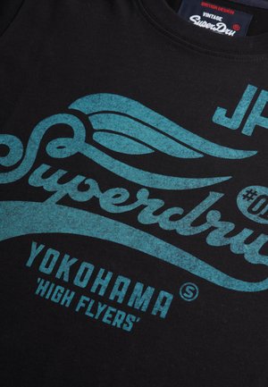 Czarna bawełniana koszulka z turkusowym, vintage'owym nadrukiem graficznym, zawierająca słowa "Superdry", "Yokohama" oraz dekoracyjne akcenty w postaci spirali.