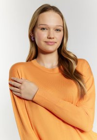 Leichter orangefarbener Pullover mit Rippbündchen und Rundhalsausschnitt, der eine glatte Textur aufweist. Die Person hat lange Haare und hält sanft ihren Arm.