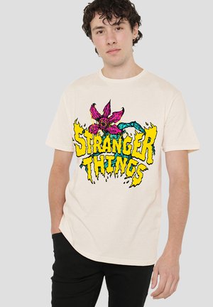 Cream-colored cotton t-shirt featuring a vibrant, graphic design with a pink flower and "STRANGER THINGS" text in yellow and blue.