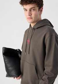 Felpa grigia in cotone con tasca frontale, con un logo rosso. Nella mano opposta viene tenuta una borsa a tracolla nera in pelle. Design casual.