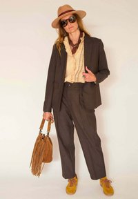 Blazer marron ajusté sur un chemisier beige à volants, pantalons gris, baskets jaunes et un sac à franges marron. Les accessoires incluent des lunettes de soleil et un chapeau.