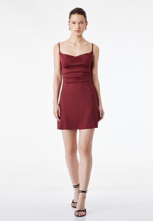 Kobieta ubrana w bezrękawnikową, sięgającą kolan bordową sukienkę z marszczeniami, czarne sandały na cienkich paskach, stojąca na tle gładkiej białej ściany.