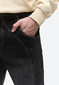 Hand i framficka på svarta jeans med orange sömmar, bär en krämfärgad sweatshirt med ribbad mudd och broderat "B" på ärmen.