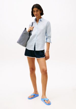 Chemise bleu clair à boutons, shorts navy avec bord blanc, tongs bleues et sac fourre-tout rayé noir et blanc. Textures lisses.