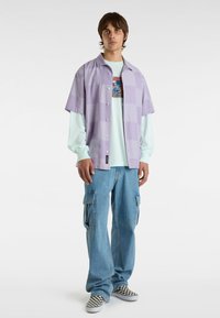 Camicetta a maniche corte a scacchi lavanda sopra una maglietta a maniche lunghe con grafica verde menta, abbinata a jeans cargo blu chiaro e scarpe a scacchi bianco e nero.