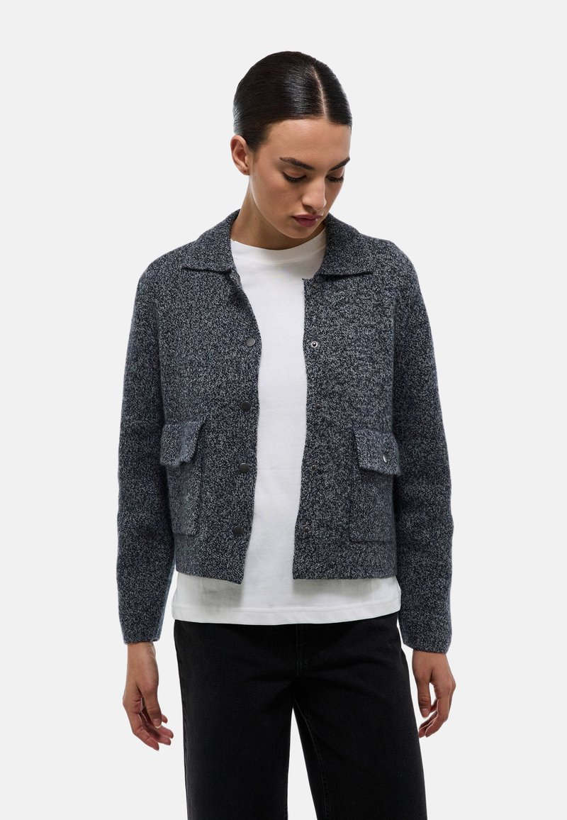 Veste tricotée grise, texturée, avec un col, des boutons sur le devant et deux poches poitrine. Portée par-dessus une chemise blanche, accompagnée de pantalons noirs.