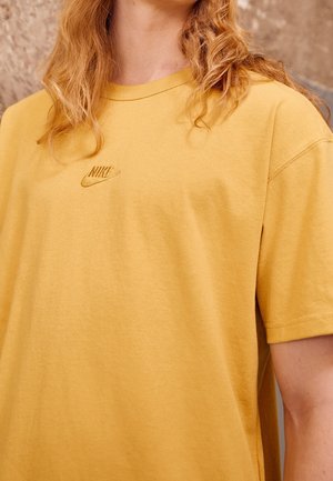 Personne aux cheveux blonds ondulés portant un t-shirt jaune Nike avec un petit logo brodé sur la poitrine.