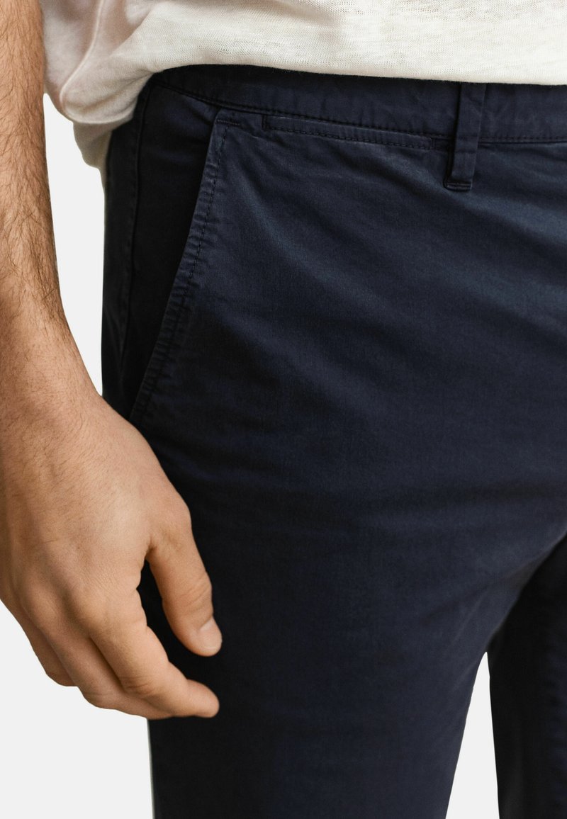 Mano de hombre apoyada junto al bolsillo de unos pantalones azul marino, con una camisa beige clara metida por dentro de la cintura.