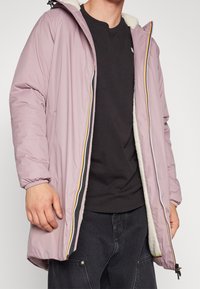 Personne portant une veste à capuche violet clair avec doublure en polaire blanche, dézippée sur un T-shirt noir uni et un pantalon foncé.