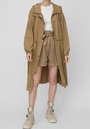 Persoon draagt een lange beige kapmantel over bijpassende korte broek met hoge taille, wit shirt en beige veterlaarzen tot aan de enkels.