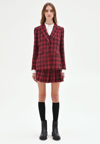 Completo a quadri rossi e neri con blazer su misura e gonna a pieghe. Presenta una camicia bianca con colletto sotto e stivali neri alti fino al ginocchio.