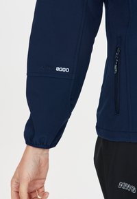 Mörkblå vattentät jacka med W-PRO 8000-märke. Har en ficka med dragkedja, elastiska ärmslut och en slät yta. Närbild av ärmen.