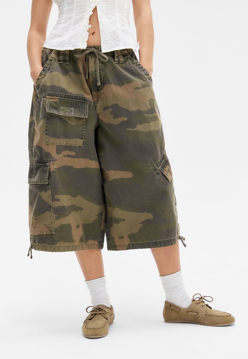 Calções cargo camuflados em verde e castanho, com um corte descontraído, cintura com cordão e múltiplos bolsos. Tecido texturizado com punhos elásticos.
