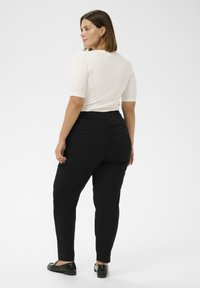 Sieviete ar īsiem brūniem matiem, valkājot baltu īsām piedurknēm augšu, melnas augstu vidukli bikses un melnus loaferus, stāv un skatās sānis.