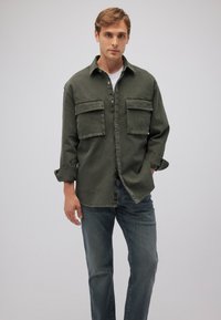 Dunkelgrüne Denim-Hemd mit zwei Fronttaschen, Knopfleiste und hochgerollten Ärmeln, kombiniert mit blauen Jeans. Schlichter Hintergrund.